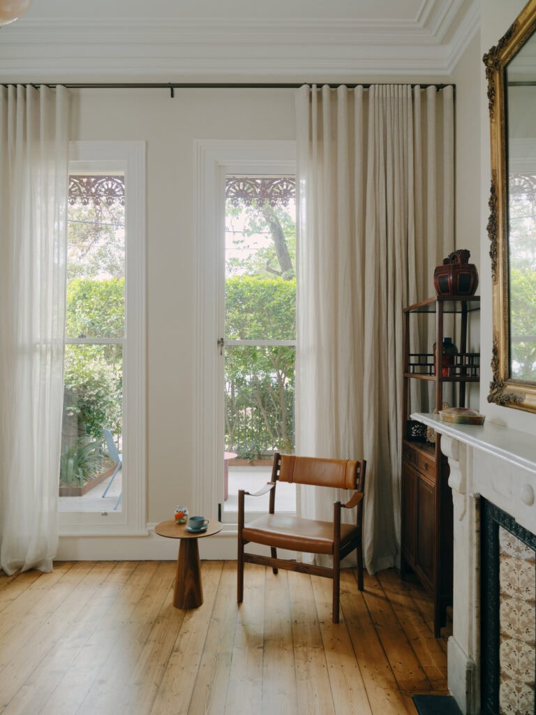 Bright living room with large windows, cream curtains, and wooden furniture.